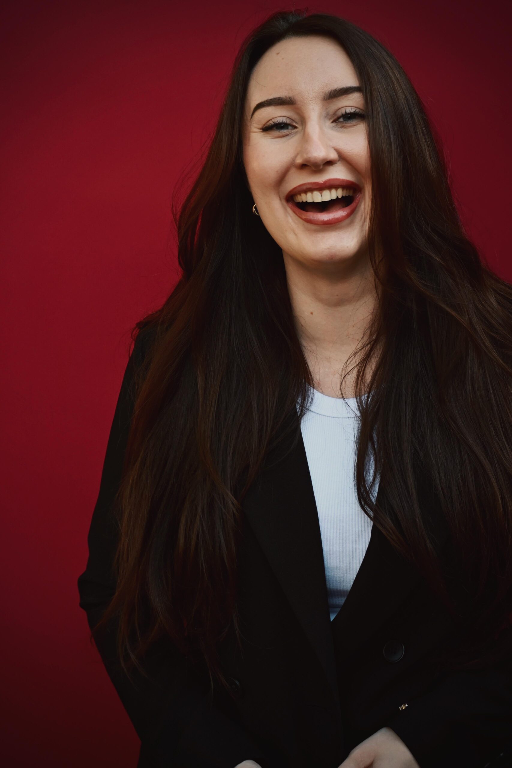 Kim Denie, jonge witte vrouw met lang bruin haar, lacht, zwarte blazer met wit shirt eronder tegen een dieprode achtergrond.