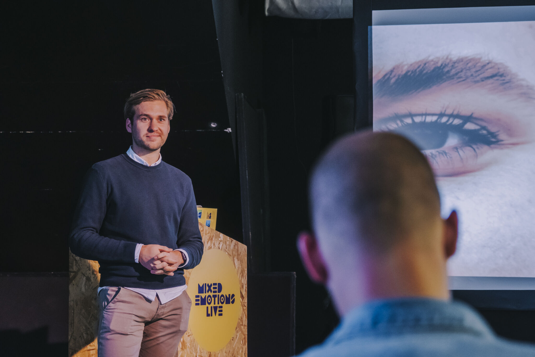 Ambassadeur Marijn van der Laan, jonge blanke man met kort blond haar draagt een donkerblauwe trui,, een lichtblauw overhemd eronder op een beige casual broek. Hij geeft een presentatie en houdt zijn handen gevouwen voor zich.