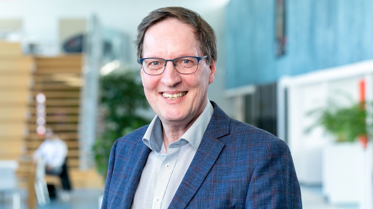 Eric heeft donkerblond haar, draagt een bril en lacht vriendelijk. Hij draagt een lichtblauw overhemd met een blauw colbert. Hij staat in een zogenoemde kantoortuin, die achtergrond is wat wazig gemaakt.