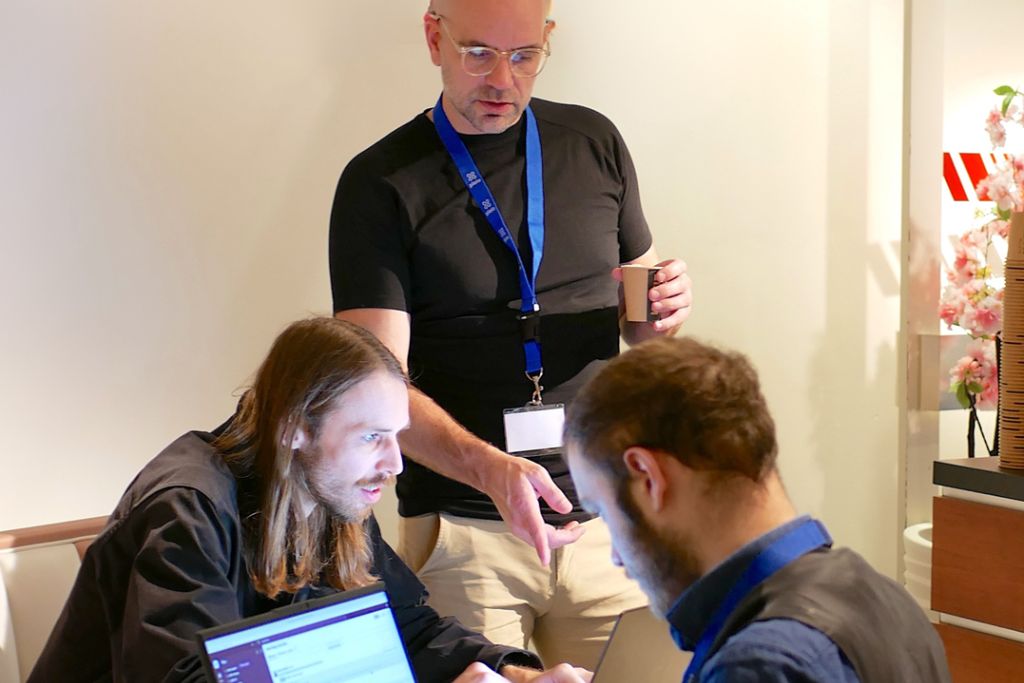 Twee mannen achter een laptop zitten tegenover elkaar. Erachter staat een derde man die deze 2 deelnemers op de deelnemersdag van Pleio, instructies geeft over Pleio's software.